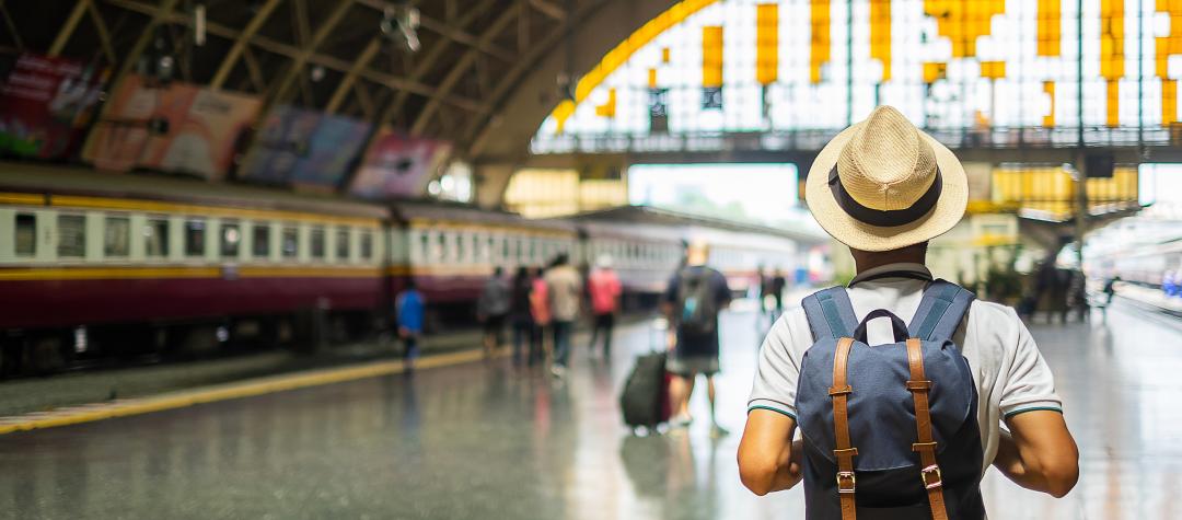 A traveler wearing a backpack and hat, walking towards a train